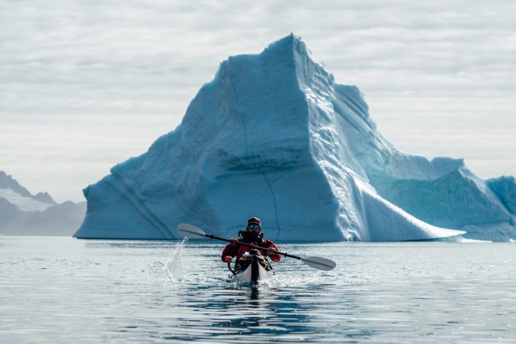 A kayaker paddles with a large iceberg directly behind him, Just North Of Kuummiut