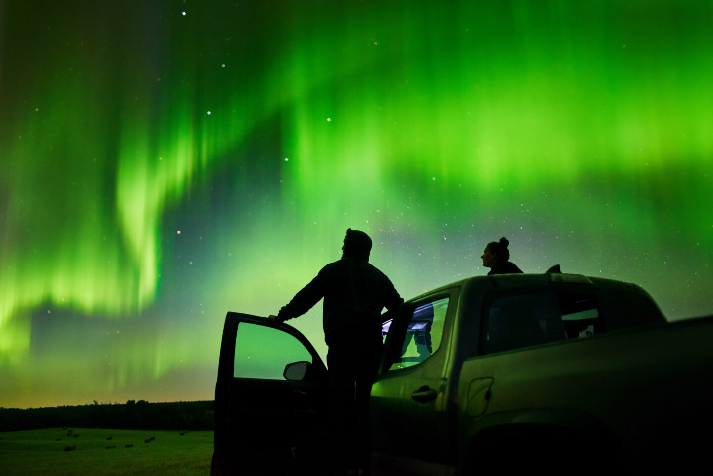 Silhouette of a man and woman standing on their pick-up truck and watching aurora borealis at night