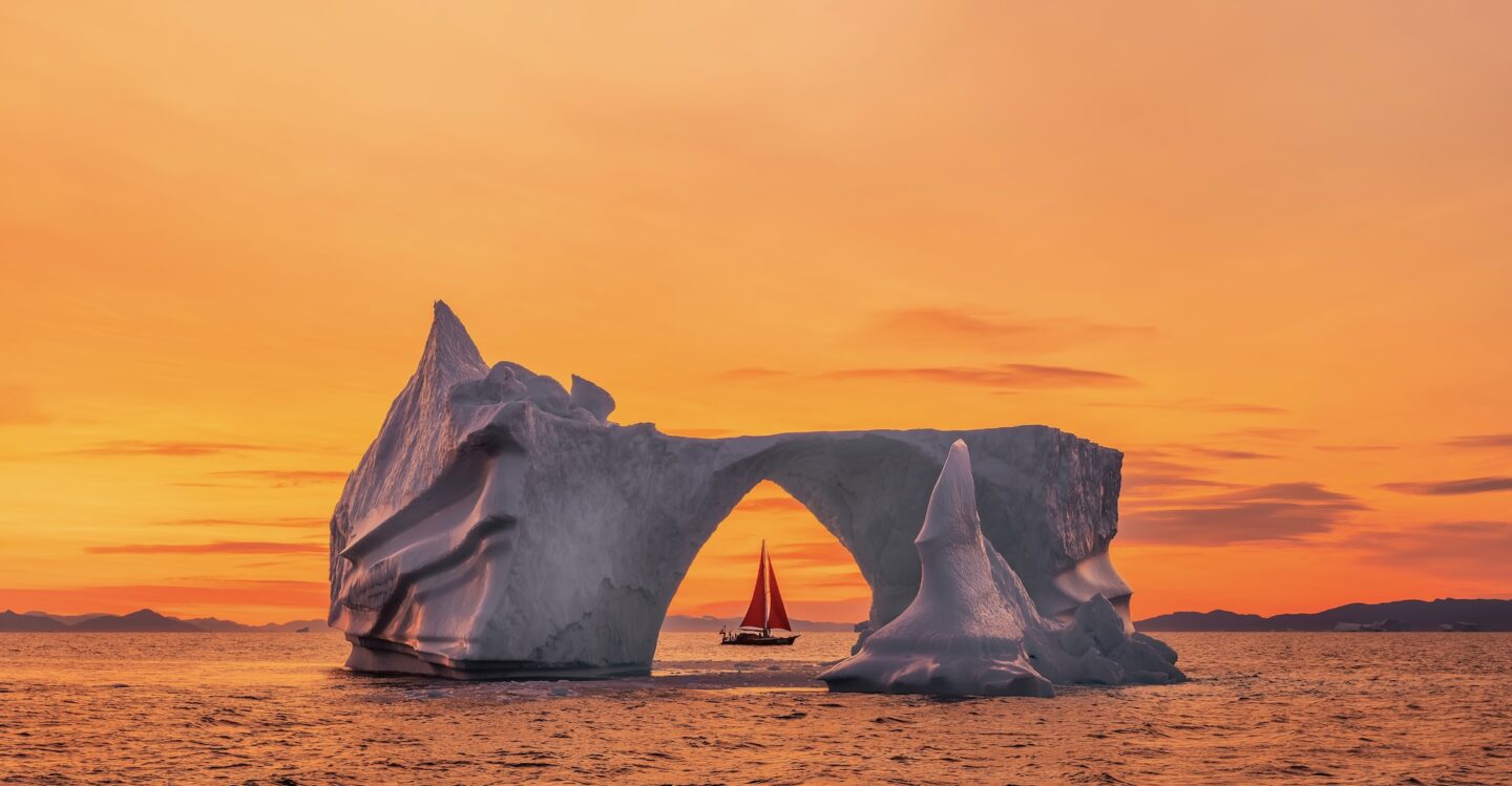 Um iceberg gigante, sol o entardecer da Groenlândia com um céu avermelhado. Ao fundo, um veleiro vermelho, apequenado diante da imensidão do iceberg.
