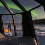 Rear view of a person seen through the glass igloo standing in the snow admiring northern lights, Lapland, Sweden