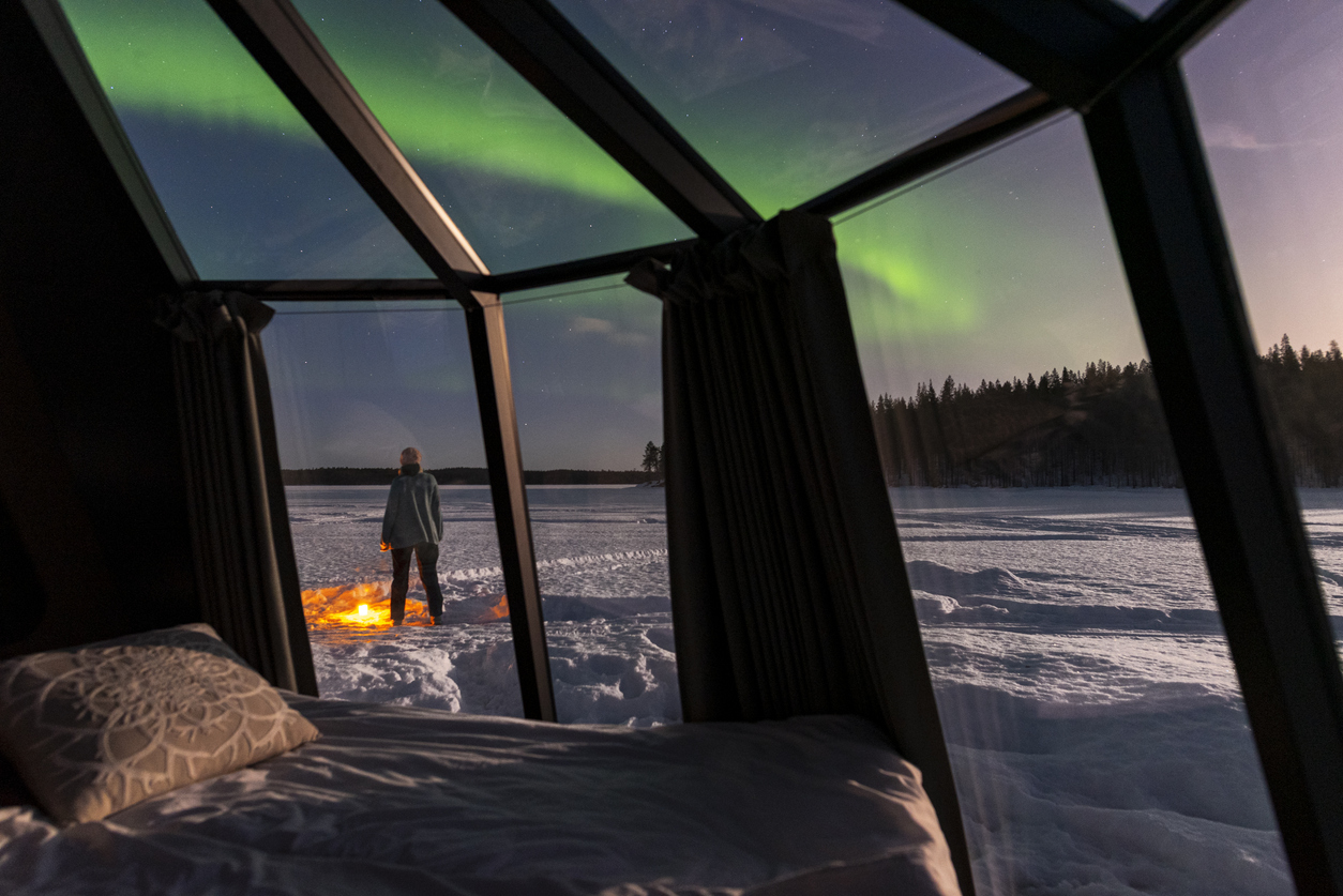 Rear view of a person seen through the glass igloo standing in the snow admiring northern lights, Lapland, Sweden