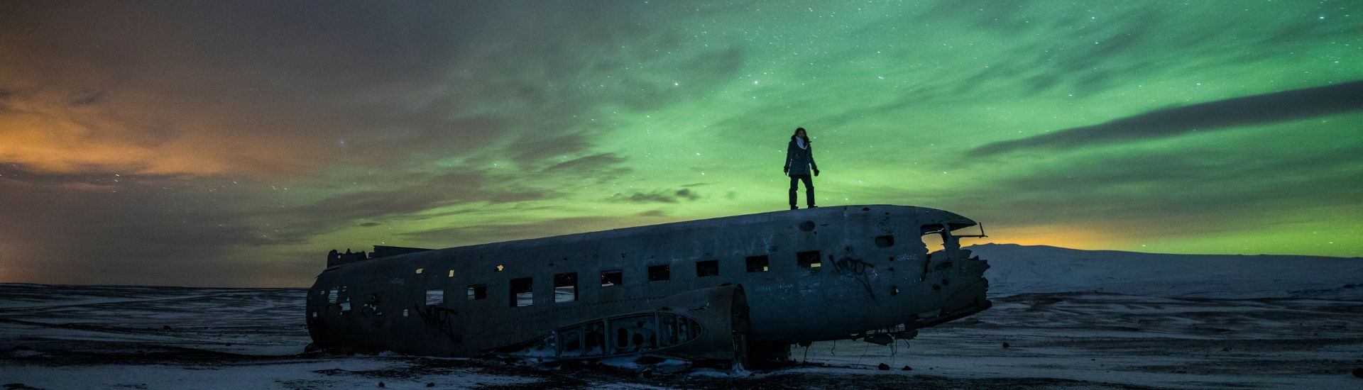 Mulher sobre os restos de um avião, na Islândia, diante da Aurora Boreal