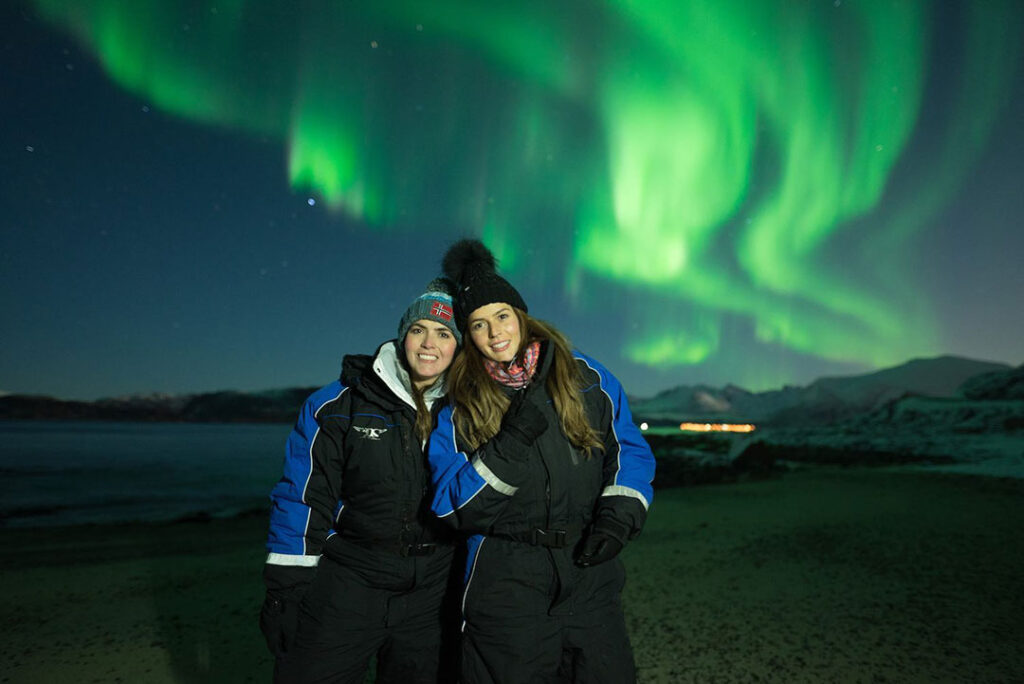 Duas mulheres posando para uma foto, com a Aurora Boreal iluminando os céus