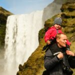 Pai carregando criança filha nos ombros em frente a cachoeira na Islândia