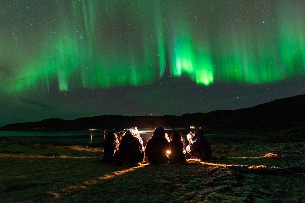 Pessoas sentadas ao redor de uma fogueira, com a Aurora Boreal em tons de verde nos céus